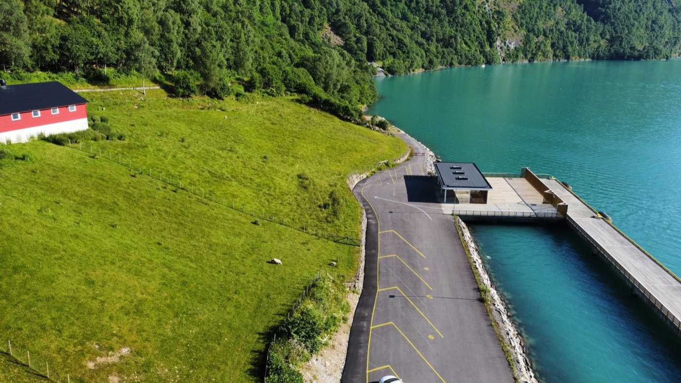The shore road approaching the cruise pier from the air, with grazing land and a red boathouse on the landward side, the marked coach bays and the terminal building with its timber deck on the other, and the pier running out into the fjord