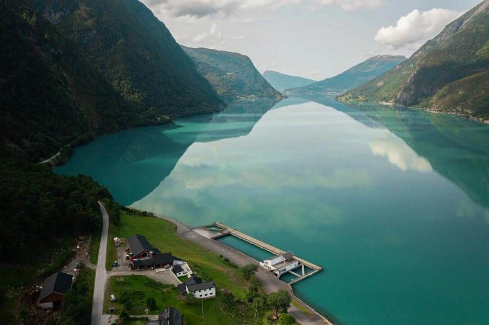 Aerial view of the cruise pier with no ship alongside: the empty quay deck, the paved coach standing area behind it, the service building and the single road that serves them, with the fjord stretching away