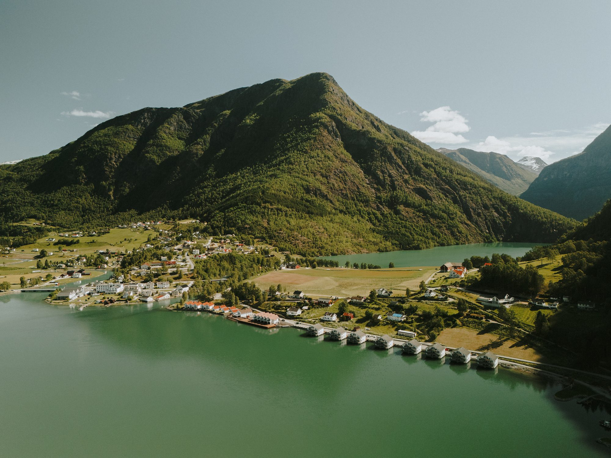 Skjolden from the air on a June afternoon: green fjord water ending at the village, a row of houses on stilts along the shore road, Eidsvatnet lying behind the fields and a forested mountain rising over it all
