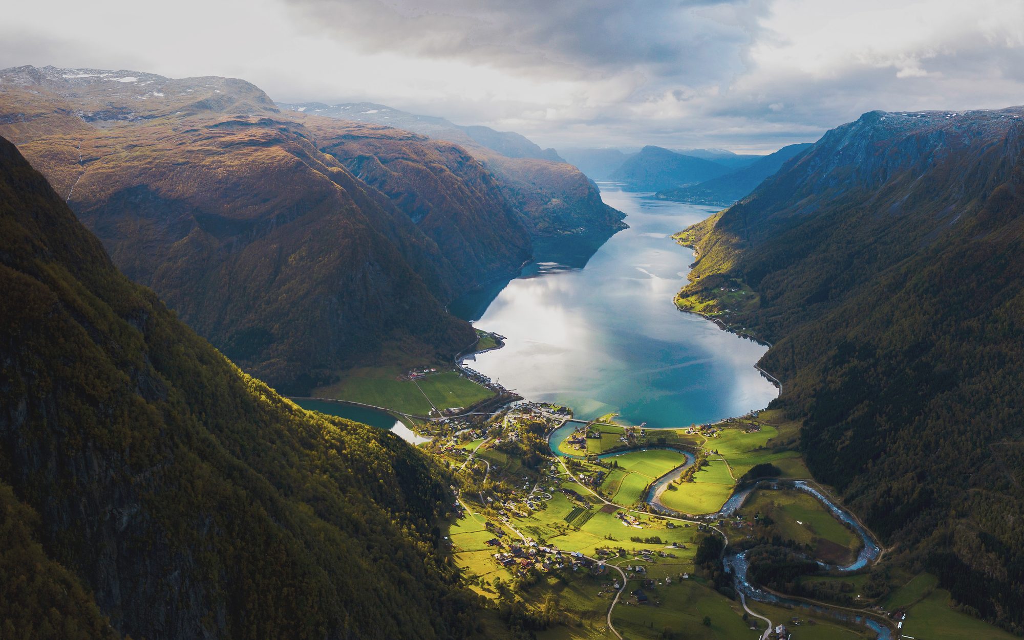 Skjolden and the head of the Lustrafjord from high above the valley: the river winding through sunlit fields to the village, and the fjord running away between steep autumn mountainsides under broken cloud