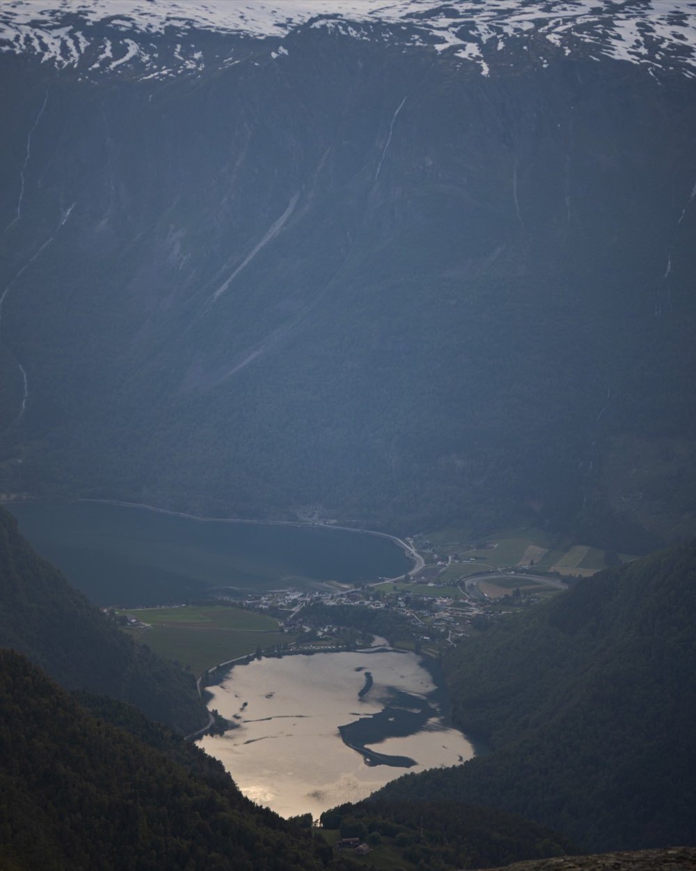 Steep view down from a summit to Skjolden, the lake and the fjord arm far below, with snowfields on the ridge opposite
