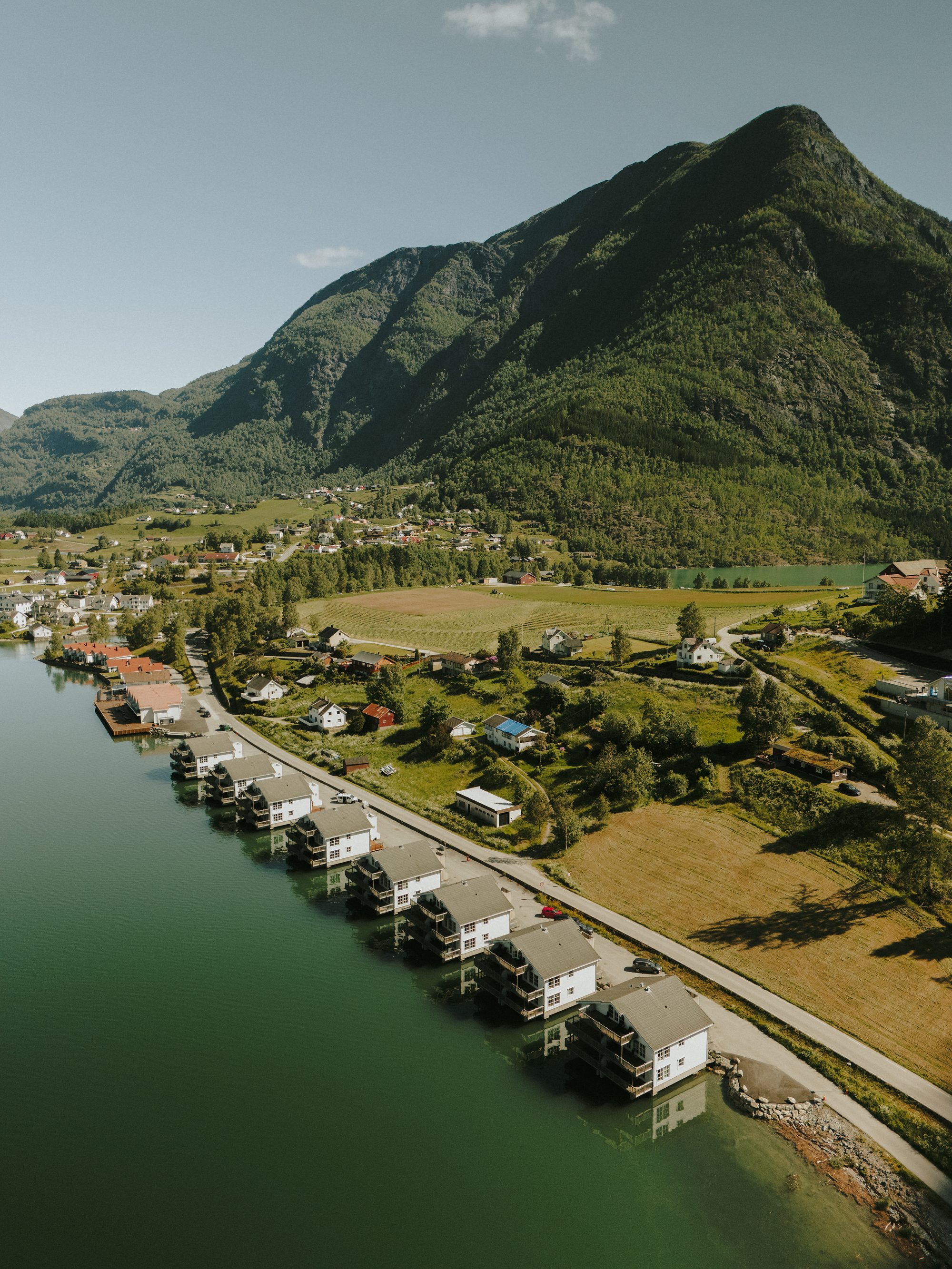 Aerial view along the shore road at Skjolden, a row of white houses standing in the green water and the village fields beneath a steep mountain