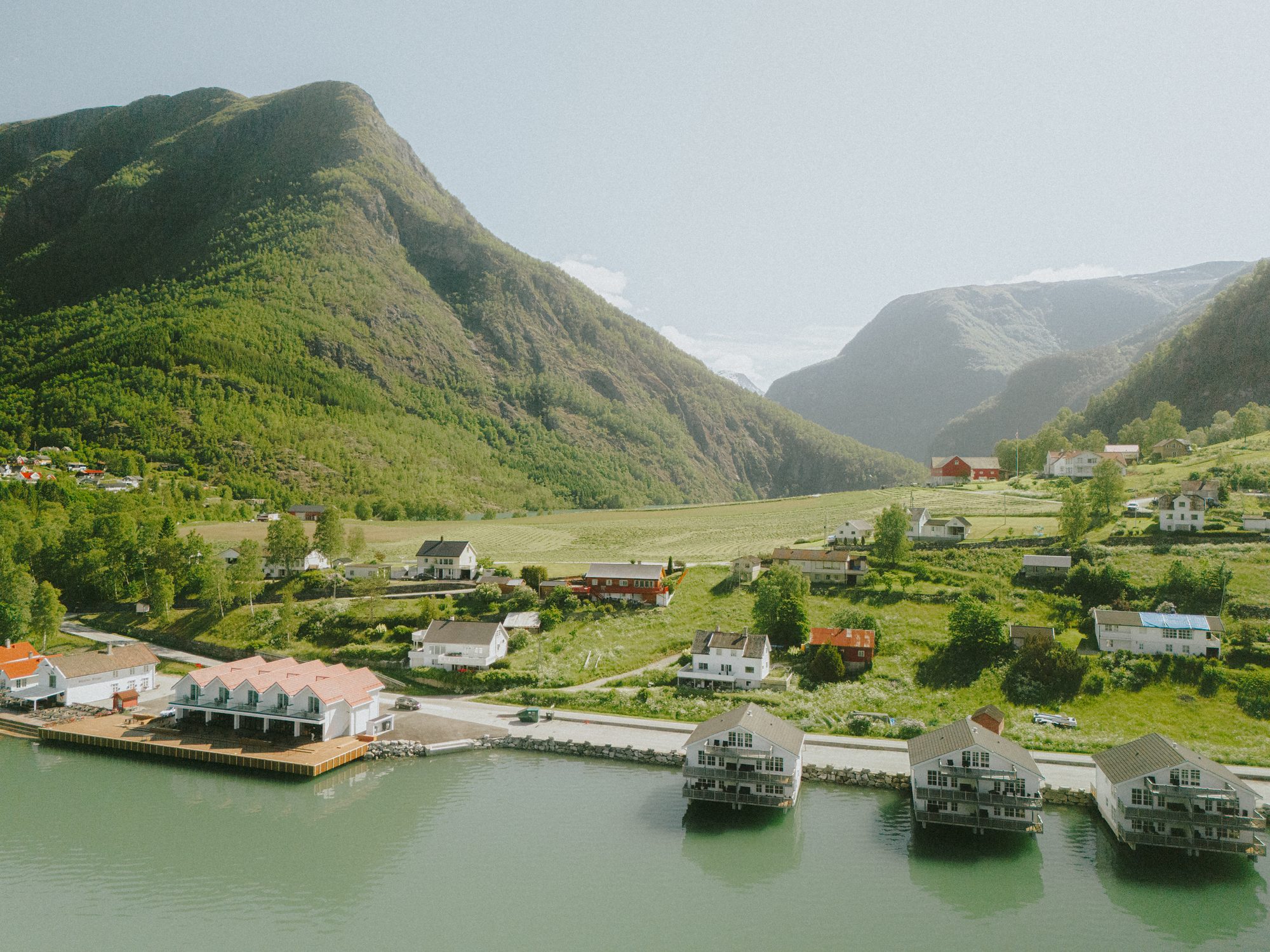 Skjolden from the air in summer: the fjord-front houses on stilts, a small pier, farmhouses among green fields and the mountains closing the valley behind