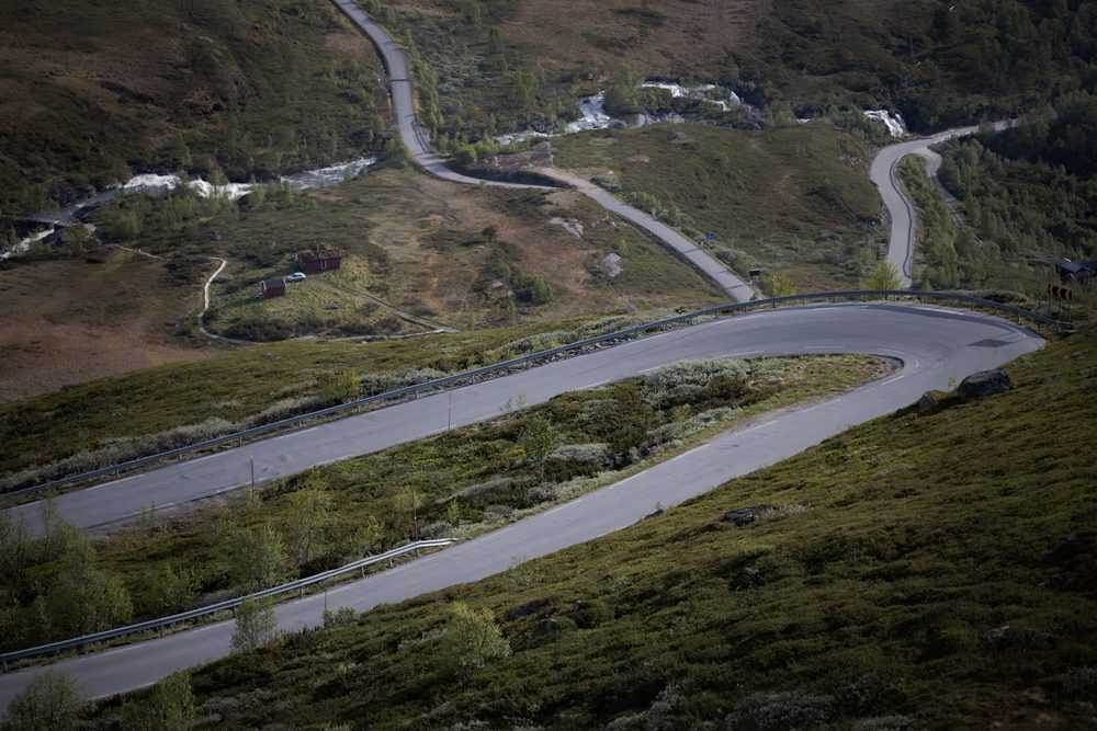 Hairpin bends of the Sognefjellet road stacked up a green mountainside, with summer huts and streams between them