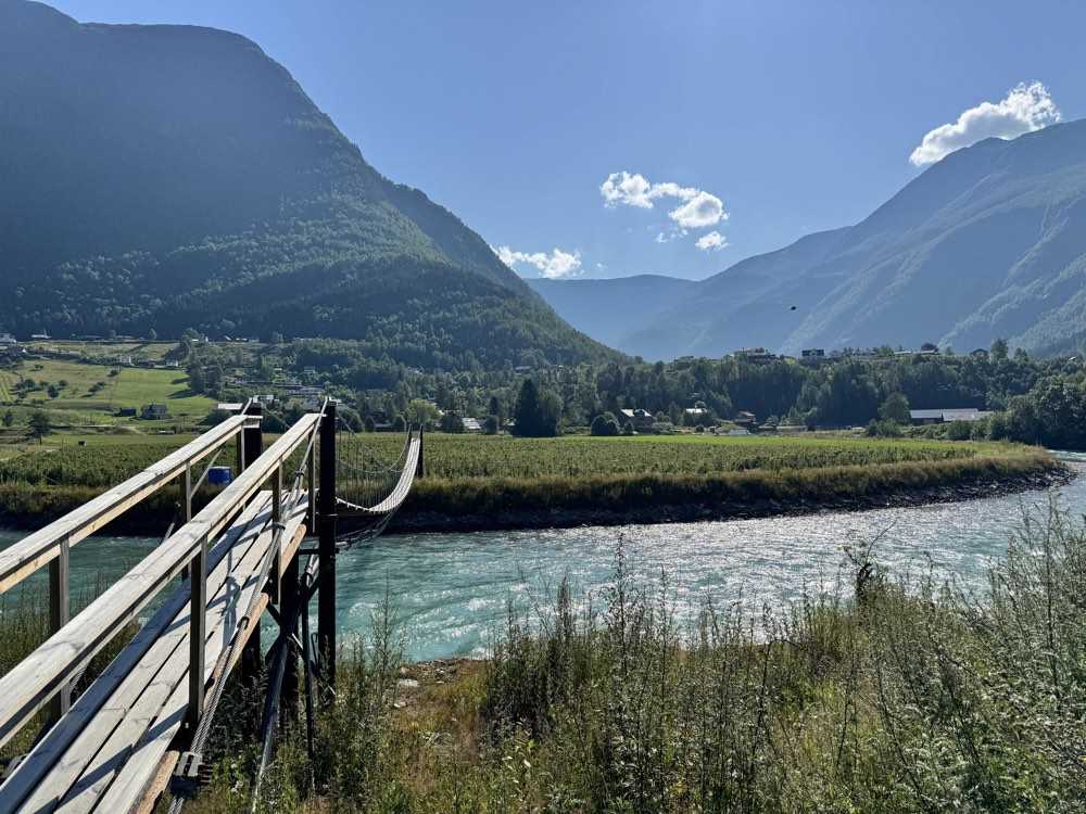 A wooden suspension footbridge over a fast turquoise glacial river, with farms, the green valley floor and mountains beyond