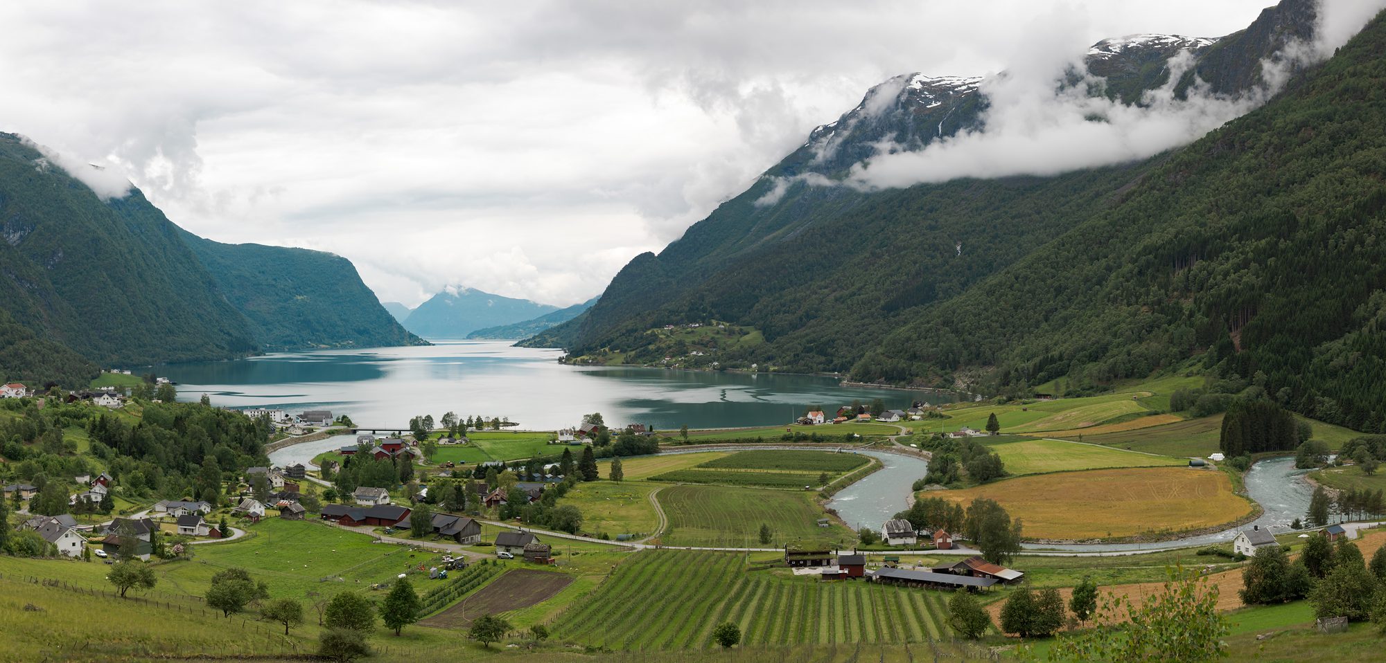 The farmed valley floor at Skjolden from above — orchards, fields and a winding river — with the fjord and cloud-topped mountains beyond