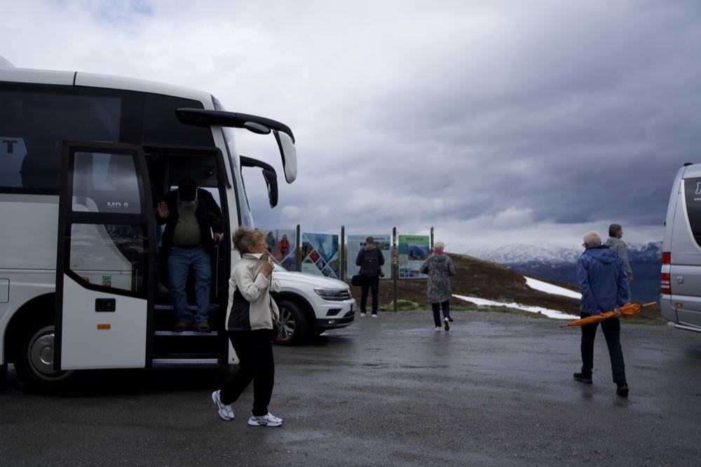 Guests stepping down from the coach at a mountain viewpoint, the MD 9 badge beside the open door, information boards and a second vehicle beyond, with snow lying on the tundra and cloud over the far ridge