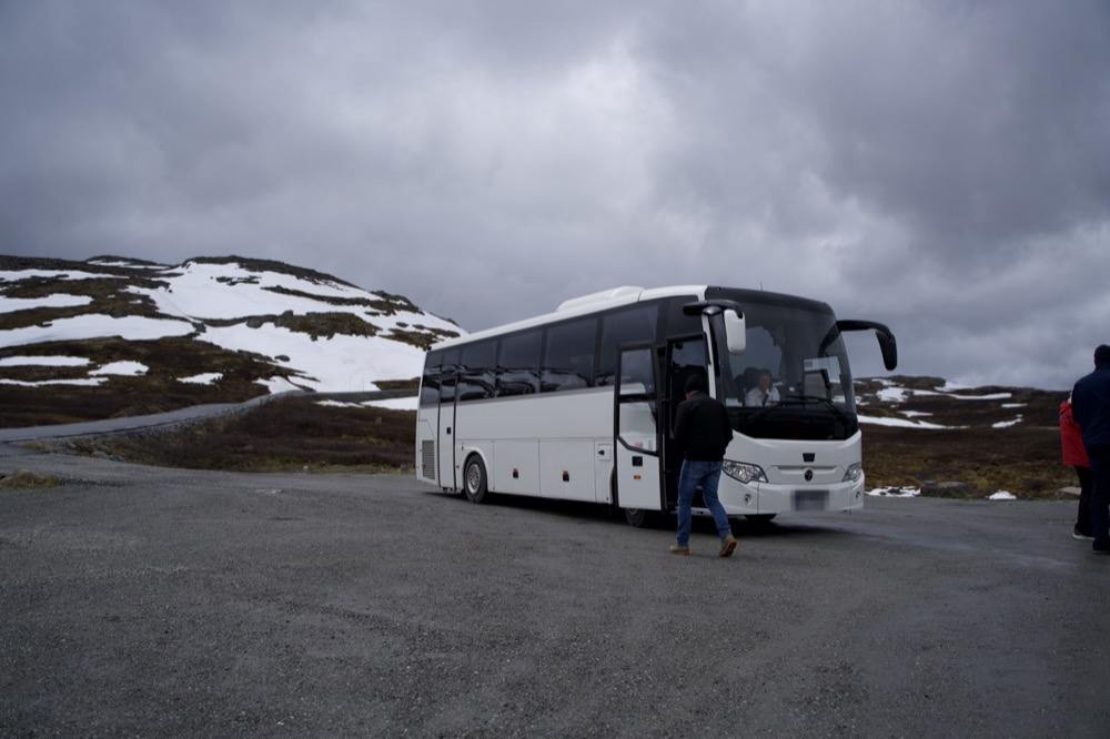 A white touring coach standing with its door open on a gravel pull-off high on the mountain pass, snow still lying in the heath around it and the road curving away into the distance under heavy cloud