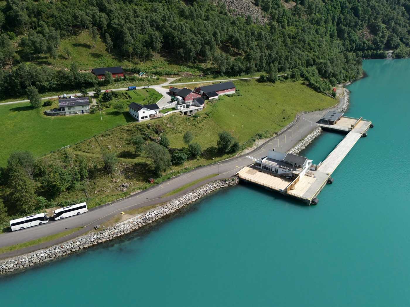 Two white coaches parked on the shore road above the fjord seen from the air, with the cruise pier and its two buildings further out on the point, farms and pasture on the slope behind and turquoise water on both sides