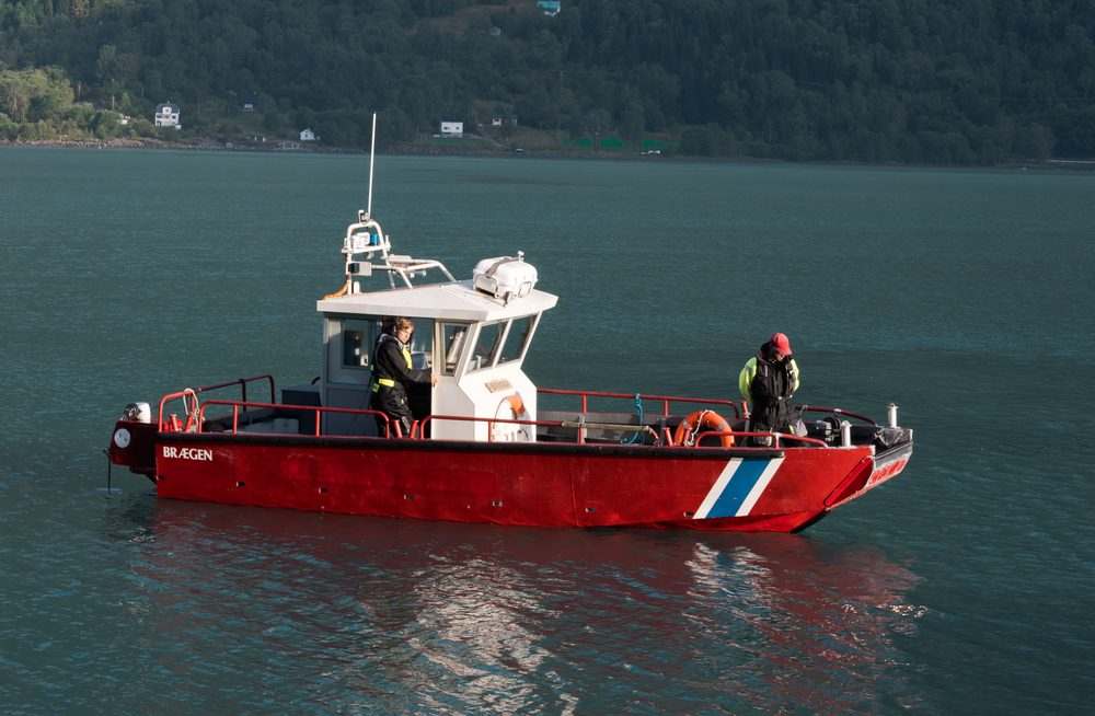 The red mooring workboat under way on the fjord with two crew in high-visibility gear on deck