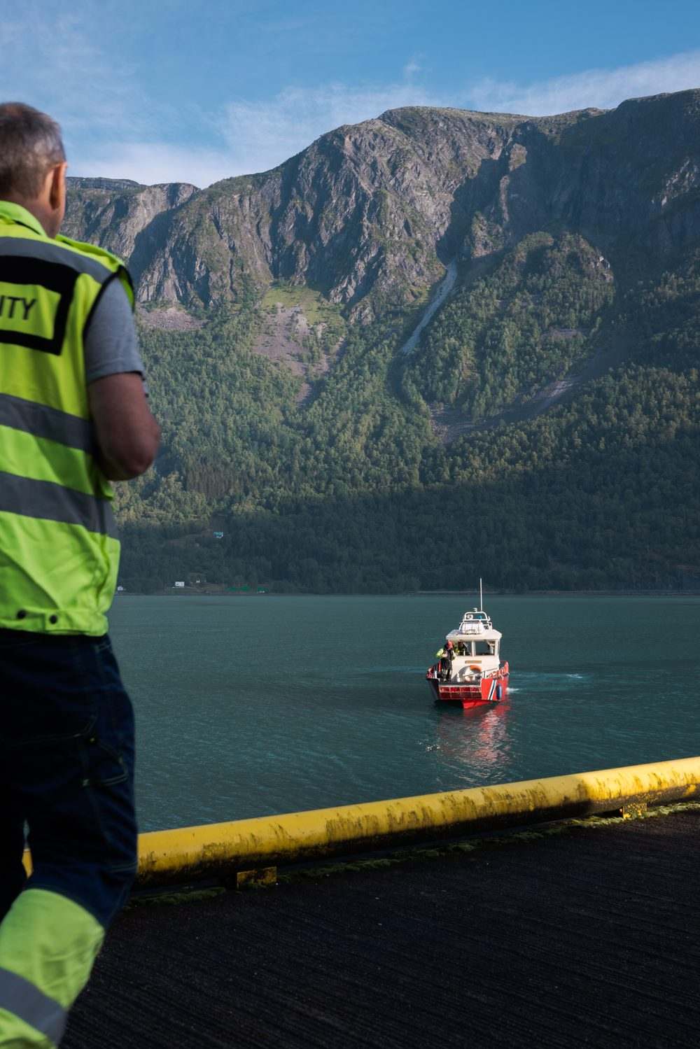 A port officer in a high-visibility security vest standing at the pier edge behind the painted yellow safety line, watching the red mooring boat come in across the fjord