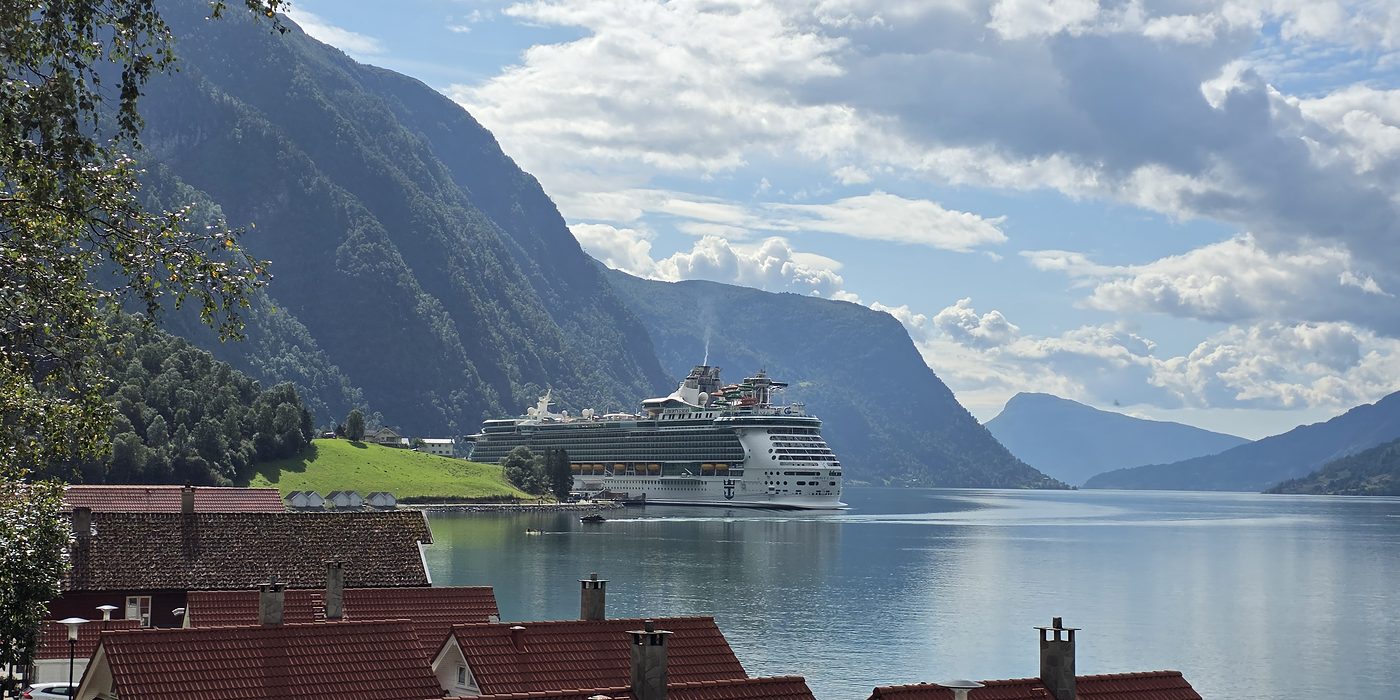 A cruise ship alongside at Skjolden in summer sunshine, seen from the village: steep green fjord walls on both sides, the Lustrafjord running out towards blue mountains, and red-tiled rooftops in the foreground