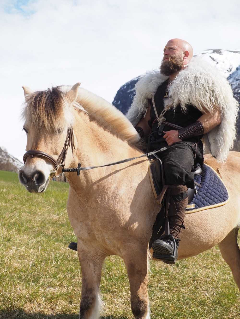 A re-enactor in a fur cape riding a Norwegian Fjord horse across a spring field below snow-covered mountains
