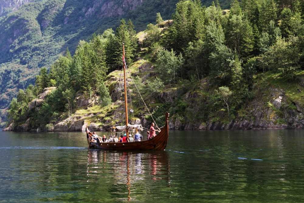 A replica Viking longship with its crew aboard and mast raised, moving across green fjord water below a steep wooded shore