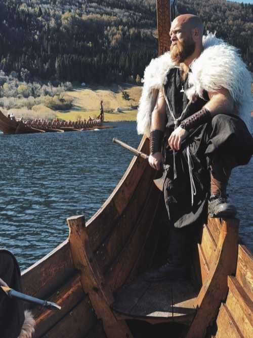 A re-enactor in a fur cape standing in the bow of a longship, with a second longship rowing behind