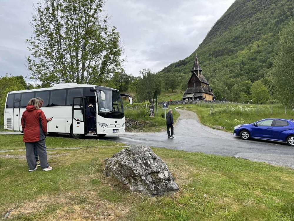 A tour coach parked on the grass at Urnes stave church as guests step off and walk up toward the church