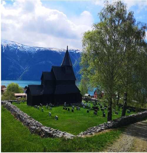 Urnes stave church behind its dry-stone churchyard wall and gravestones, with the Lustrafjord and snow-capped mountains behind
