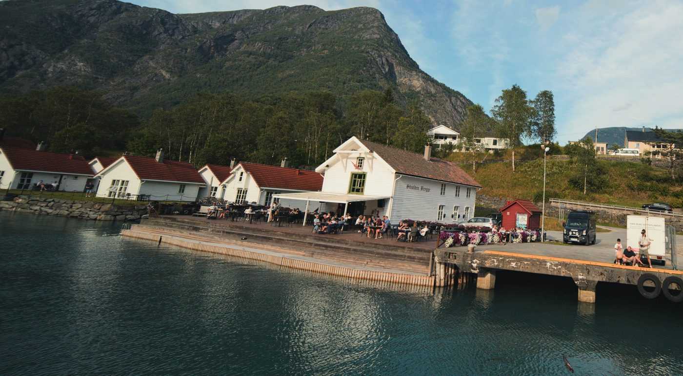 Aerial view of the quayside restaurant on a summer afternoon, its terrace tables full of guests under an awning, flower boxes along the edge and the timber wharf and red-roofed boathouses running along the shore below a steep wooded mountain