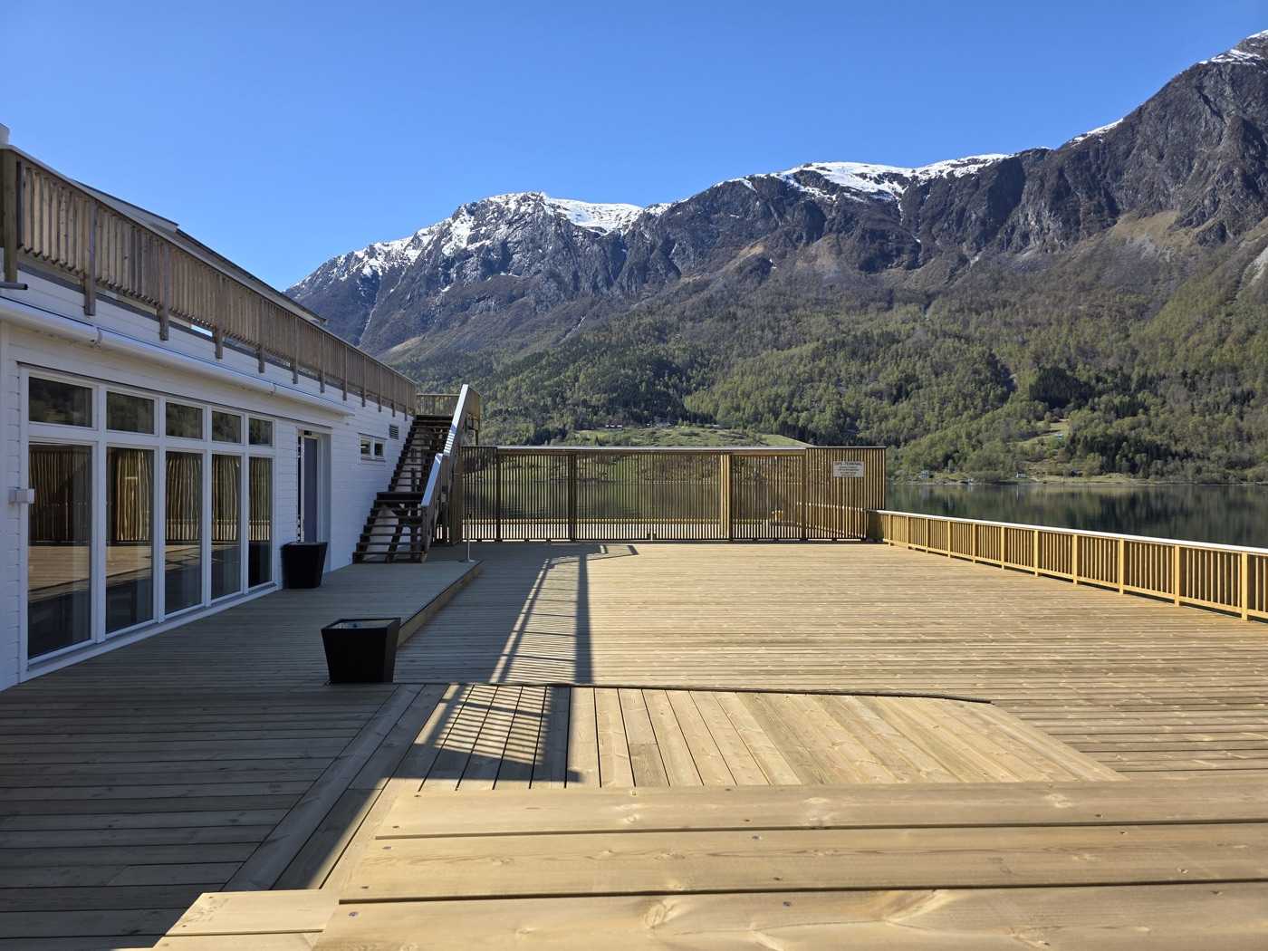 The new service building on the quay: its glazed front and first-floor balcony along the left, an external stair at the far end, and the new timber deck running out to a railing above the fjord with a snow-streaked mountain opposite