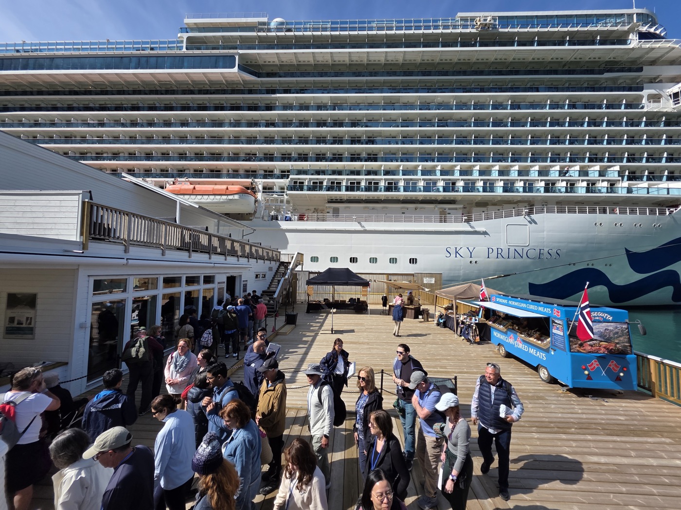 Sky Princess towering over the quay deck in sunshine, with a blue cart selling Norwegian cured meats trading beneath her bow and guests moving across the timber deck