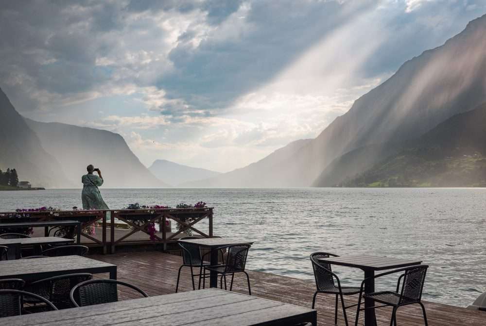 A woman photographing sunbeams over the fjord from the quayside terrace after the ship has gone, the tables around her empty