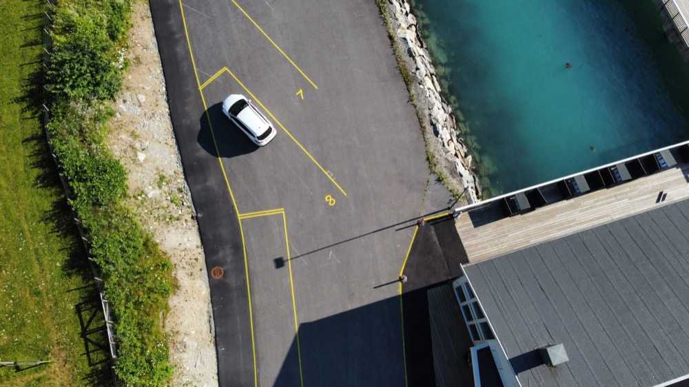 Straight-down view of coach bays 7 and 8 outlined in yellow on the apron, a car standing in one of them, and the barrier across the access road at the point where it reaches the terminal building and the timber deck