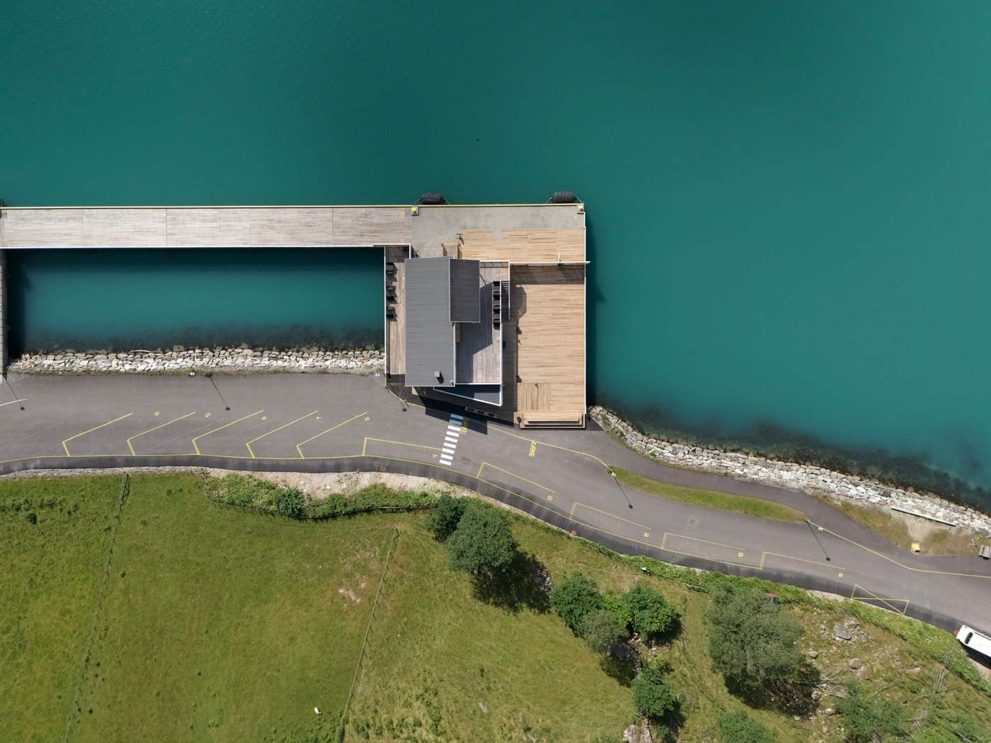 The pier and the coach standing area in one frame from directly above: the deck reaching out over the water with the terminal building at its inshore end, angled bays marked in yellow along the apron, and the linear bays continuing south past the drop zones