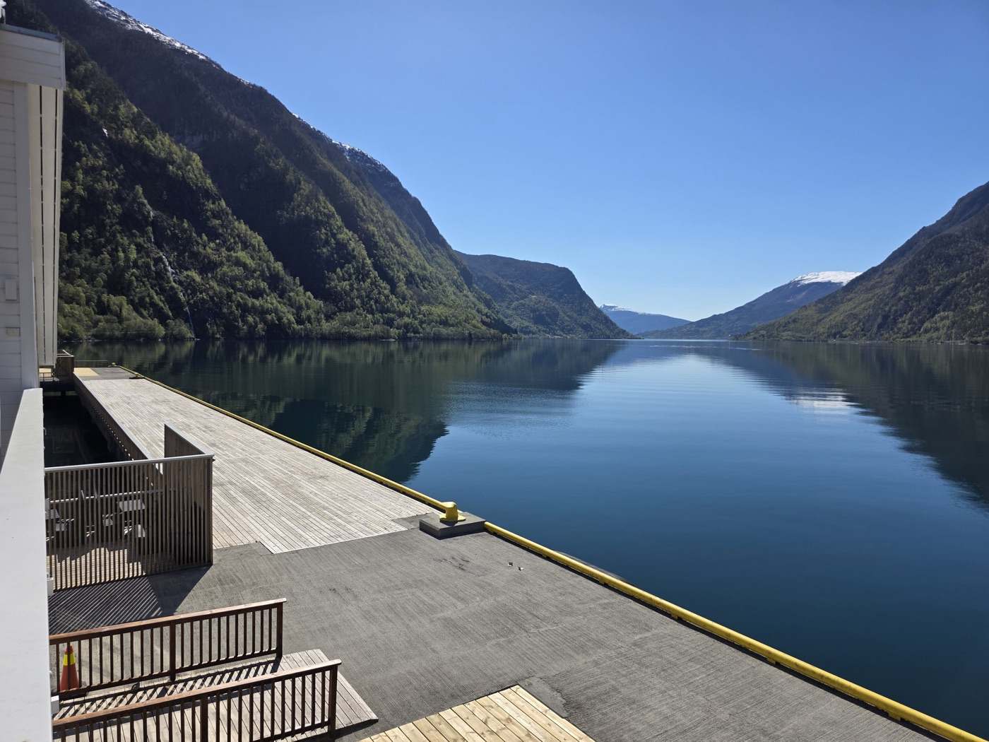 The quay on a still morning: the timber deck and the yellow-painted quay edge with a mooring bollard in the foreground, and the fjord running straight out between the mountain walls, mirror-flat under a clear sky