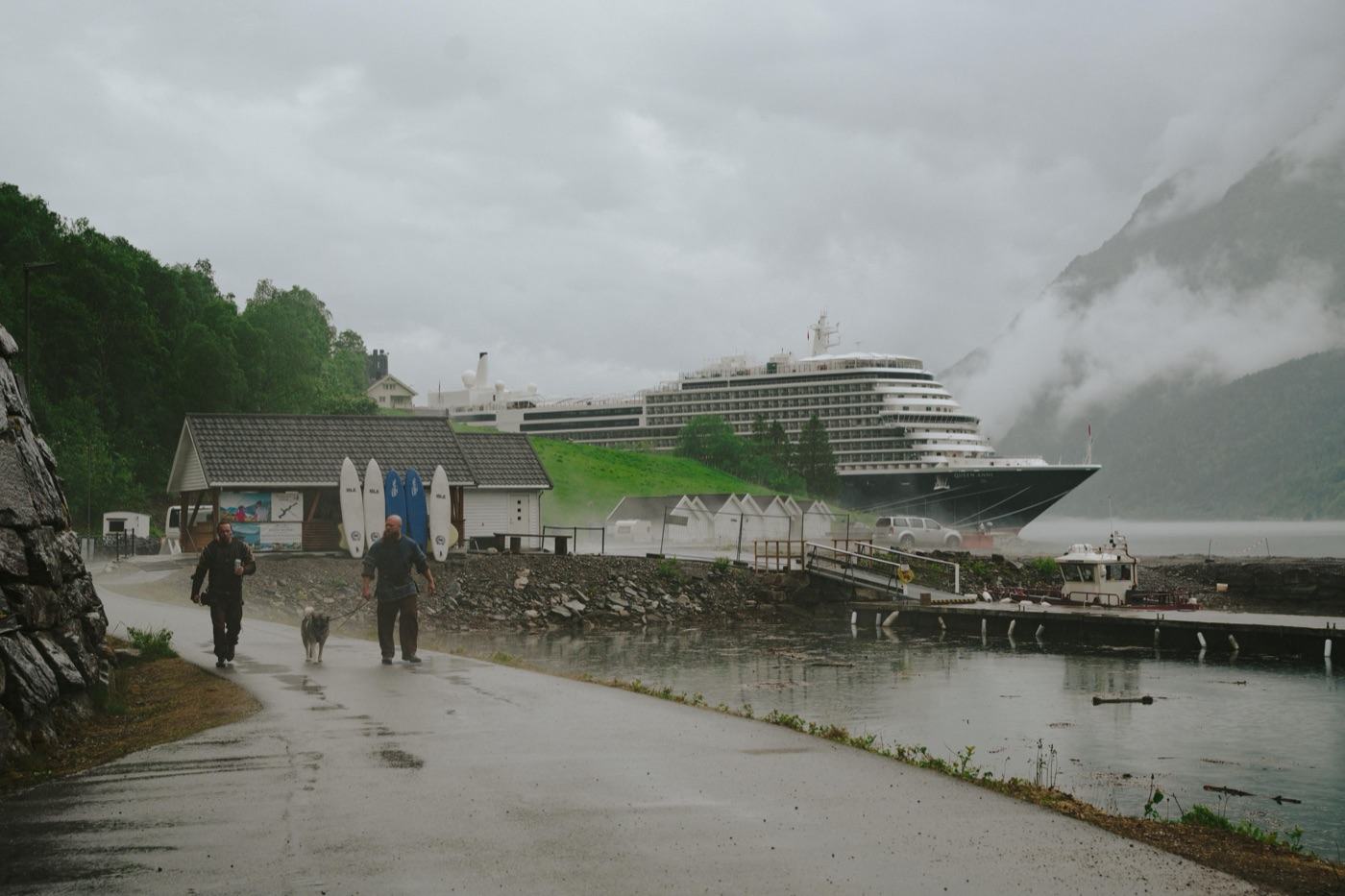Two people and a dog walking the paved shoreline path in the rain past the small white boathouses, with the cruise ship lying alongside the pier across the water and cloud halfway down the mountainside