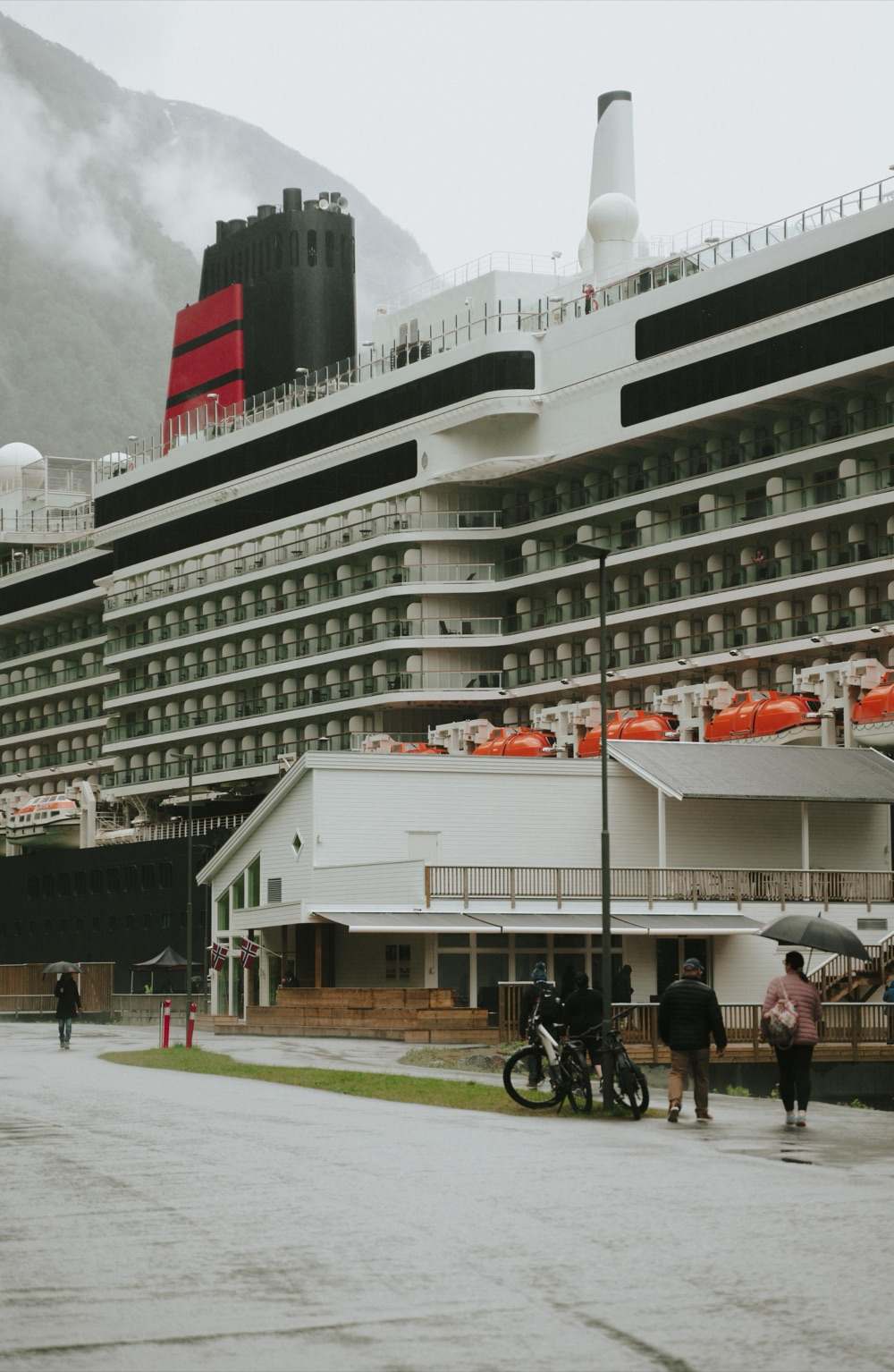 A cruise ship alongside the pier seen from the quay in the rain, her tiers of balconies and orange lifeboats rising above the white quayside building with its veranda and Norwegian flags, guests walking past two parked bicycles on the wet apron, and cloud lying in the mountainside behind