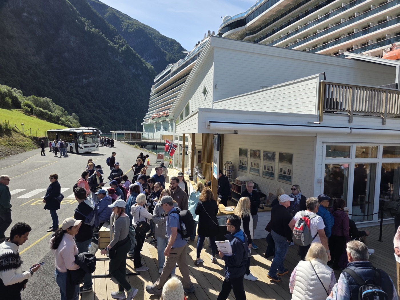 The terminal building on a sunny call day with a cruise ship rising behind it, a coach waiting on the shore road, a Norwegian flag flying, and guests filling the timber deck