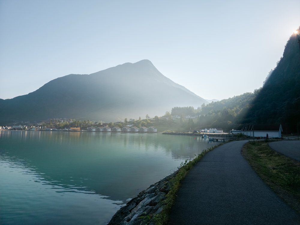 The fjord at Skjolden early on a still morning, mist lying along the mountainside and the paved shoreline walkway running past the boathouses towards the village