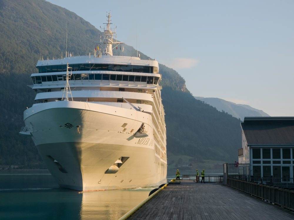 A cruise ship lying alongside the cruise pier in early morning light, seen bow-on from the quay, with three port workers in high-visibility vests on the timber deck beside her and a mooring line running ashore