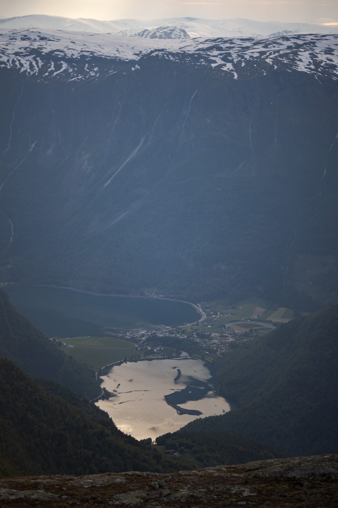 Straight down from the Berdalsnosi viewpoint: the village at the head of the fjord far below, the lake beside it catching the evening light, and snow on the mountains beyond