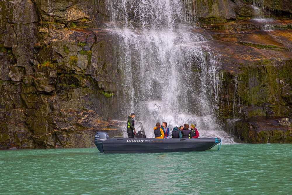 A small open boat with six guests nosed in under a waterfall sheeting down the rock face, on turquoise glacier-fed water