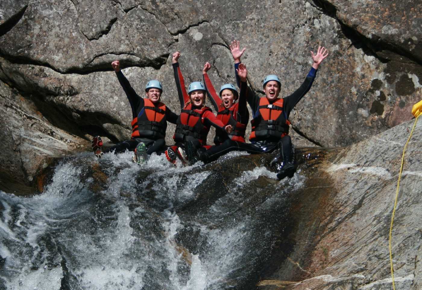 Four people in wetsuits, red buoyancy aids and pale blue helmets sitting side by side at the lip of a water-worn rock chute with their arms in the air, the stream pouring over the edge beneath them