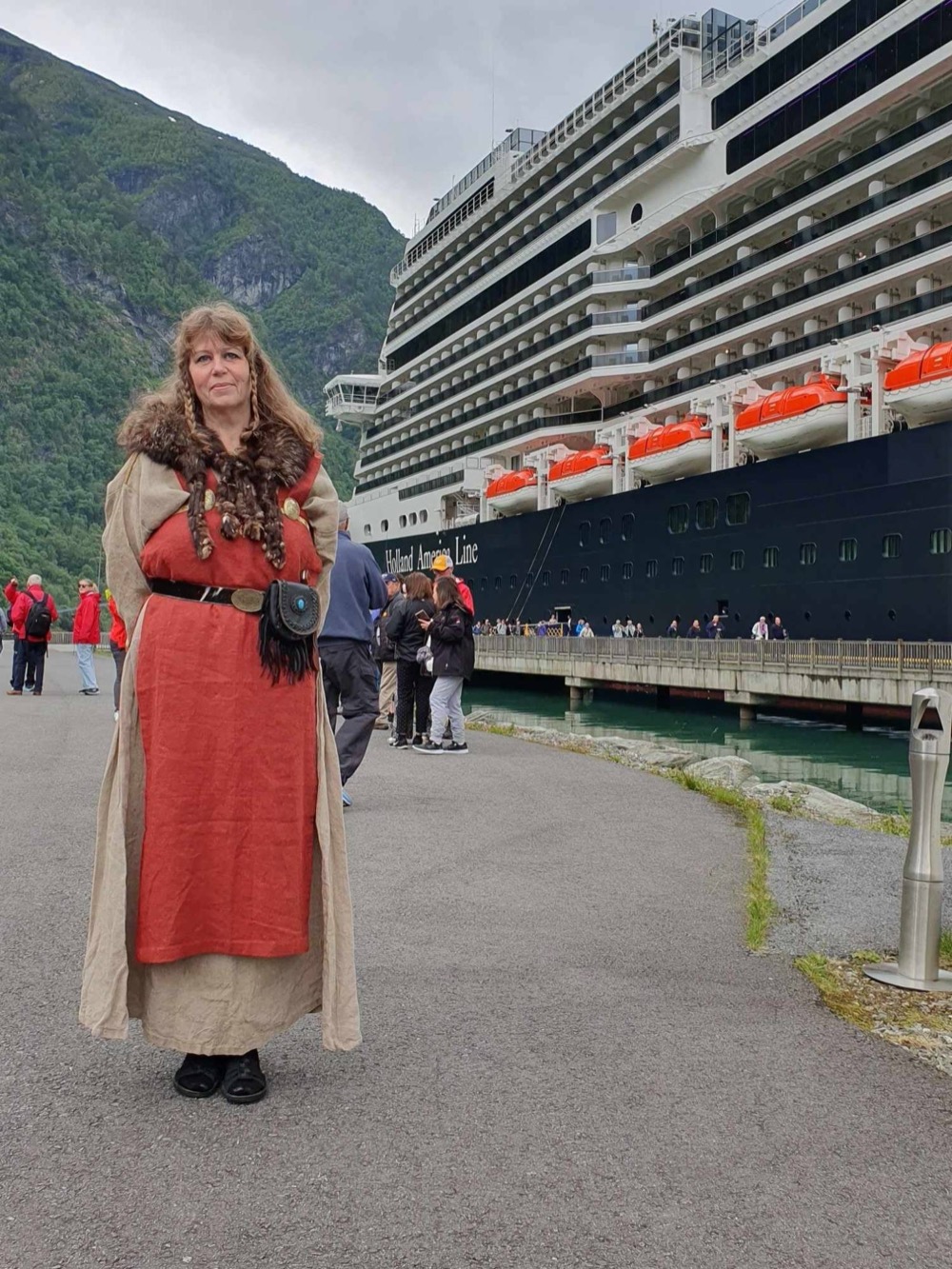 Turid Øiene of Fjordstova in Viking-age dress — a red apron dress, fur collar and braids — on a cruise quay with a ship alongside