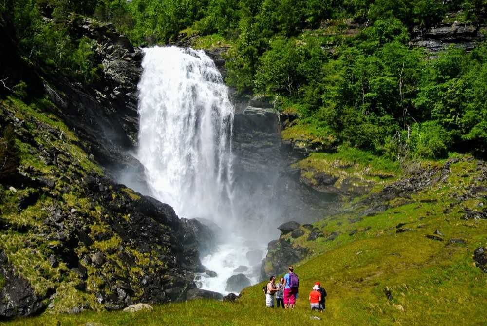 Four walkers on a green slope looking up at the Drivande waterfall pounding into its plunge pool