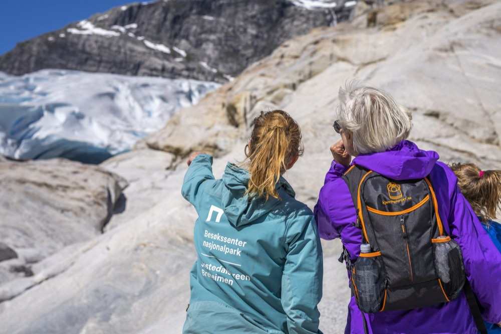 A guide in a branded jacket pointing out features of the glacier to an older visitor and a child on the ice-scoured rock