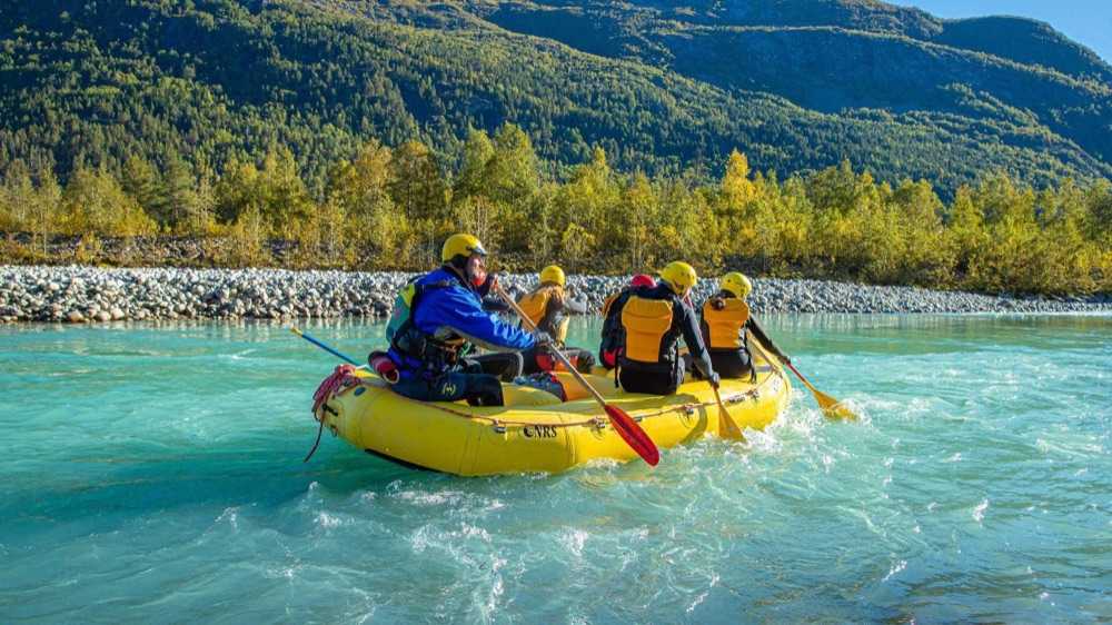 A guide and four guests in wetsuits and yellow helmets paddling a yellow raft down a wide, turquoise glacier-fed river, with a stony bank and autumn birch woods along the far side
