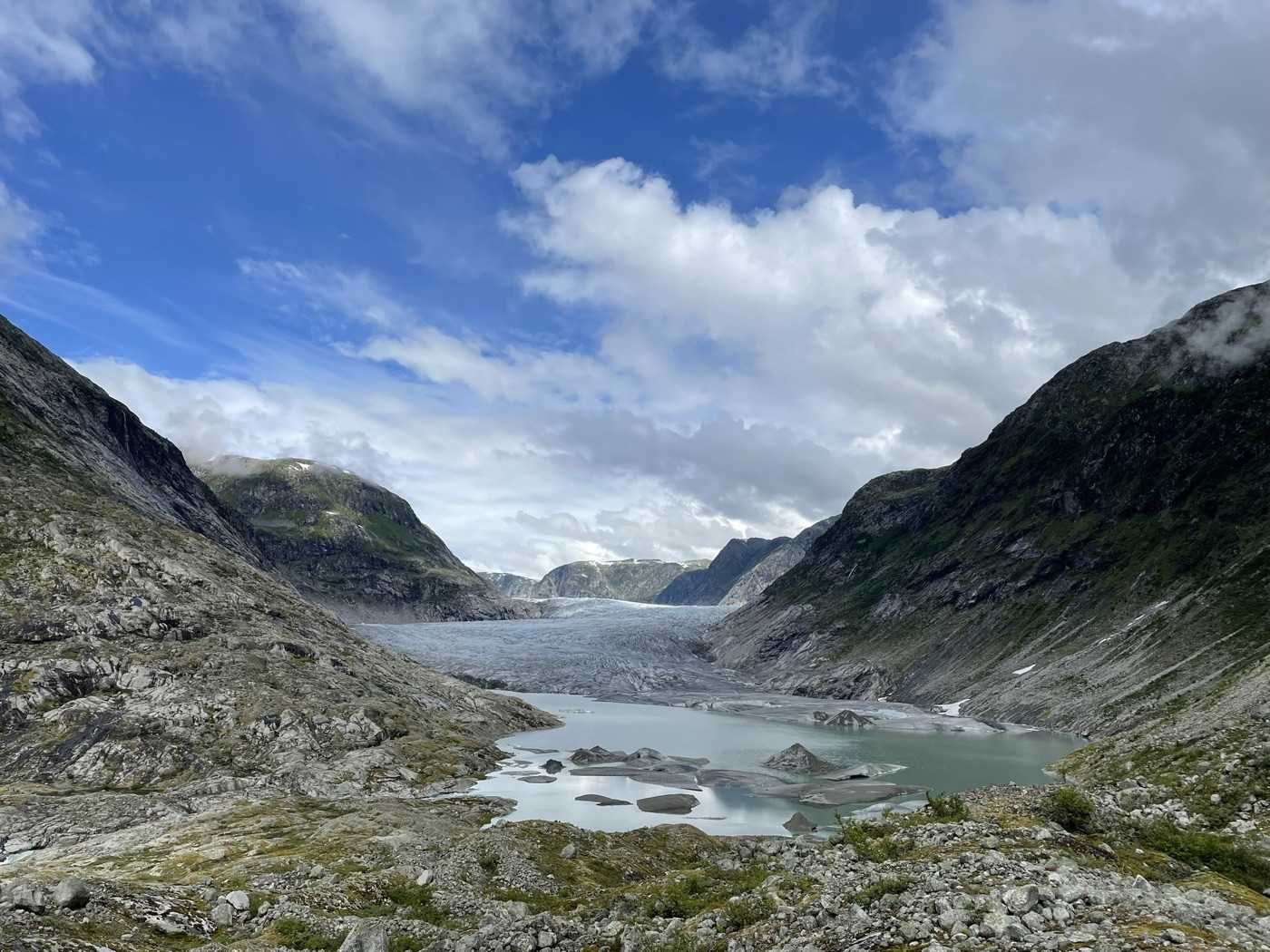 A broad glacier tongue filling the head of a rock-walled valley, its meltwater lake below it grey-green and scattered with small floes, seen across bare rock and scrub from the moraine under high cumulus cloud