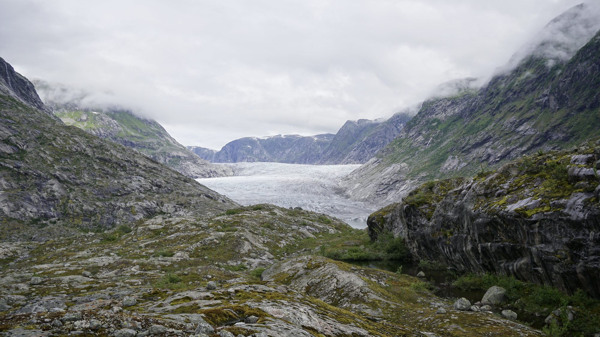 The glacier at the end of the walk: its tongue filling the head of a rock-walled valley above a meltwater lake, seen across the moraine under low cloud
