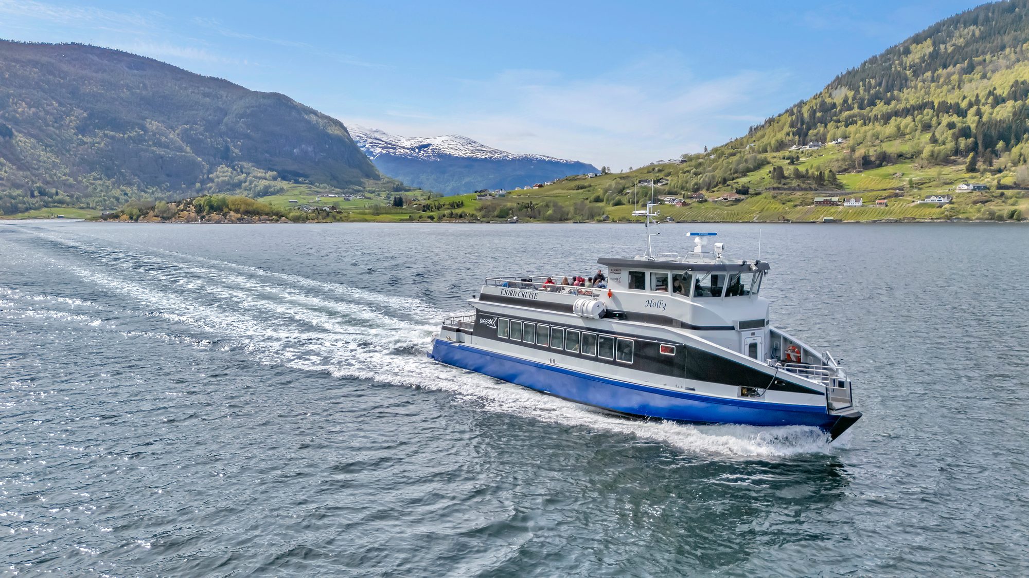 The catamaran M/S Holly running at speed on the Sognefjord, a white wake spreading behind her, guests on the open upper deck and green farmland and mountains along the far shore