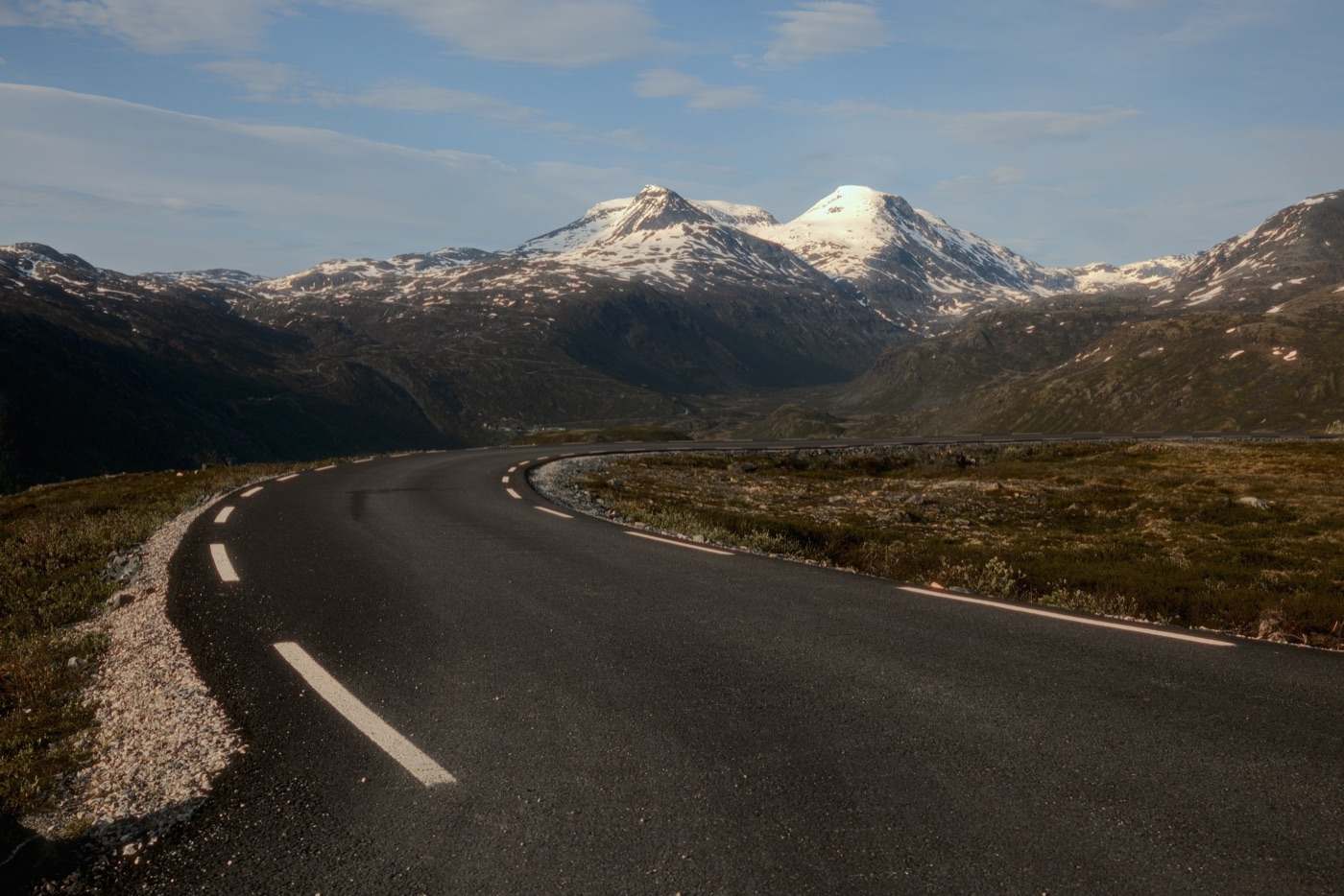 An empty mountain road curving through open tundra towards two snow-covered summits, white lines along the verge, the pass falling away in shadow between and low sun raking across the heath