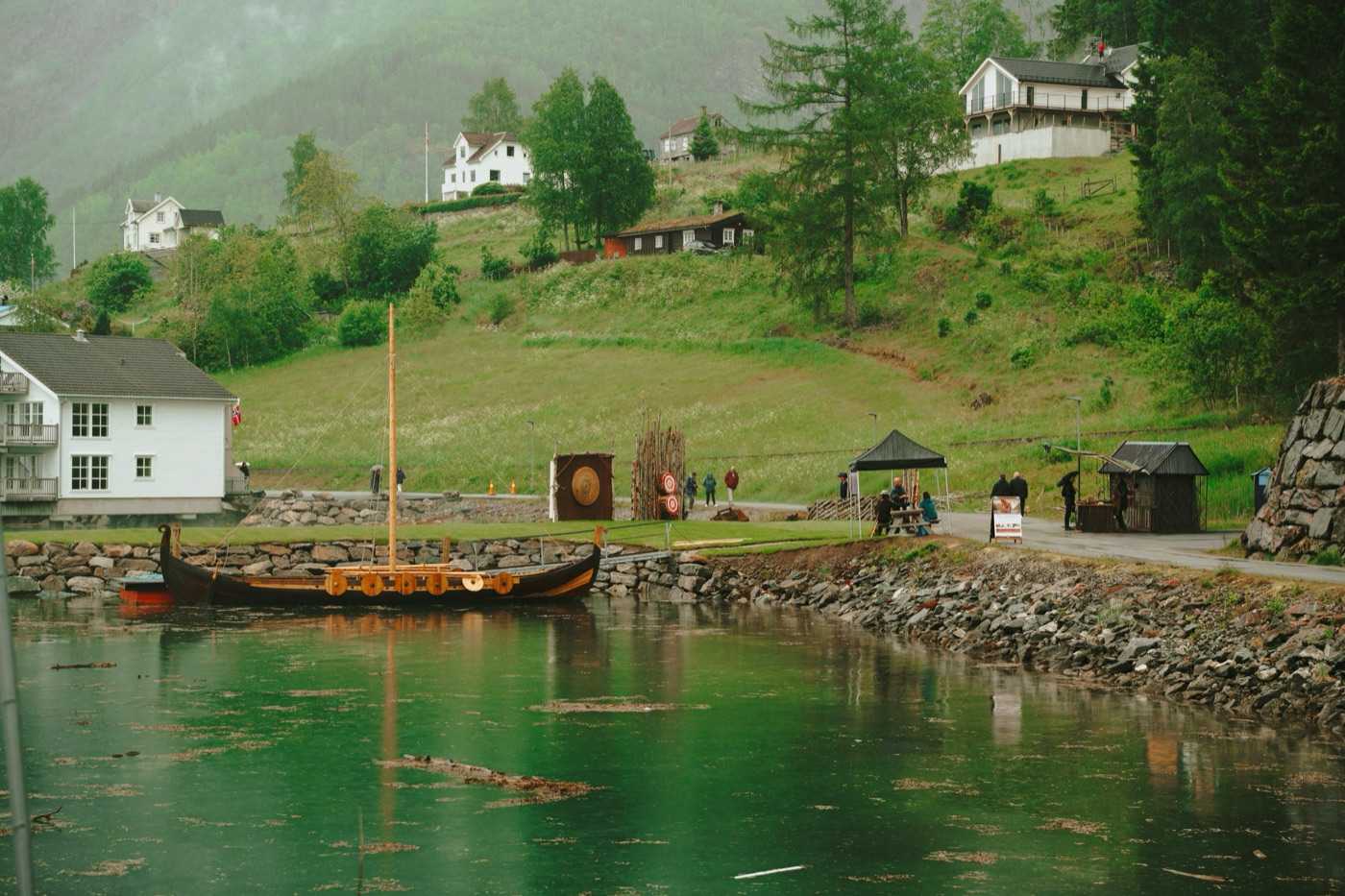 A replica Viking longship with its mast raised, moored against the stone breakwater in green fjord water, with the camp laid out on the grass behind it — a round painted shield mounted on a timber backdrop, a palisade of pointed stakes and an open shelter — and white houses on the wooded slope above