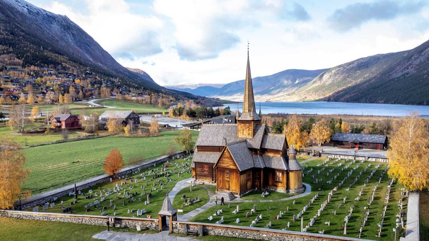 Aerial view of Lom stave church: the dark tarred timber building with its tall spire and tiered shingled roofs standing in a walled graveyard of white headstones, with golden autumn trees, the village and the river valley behind