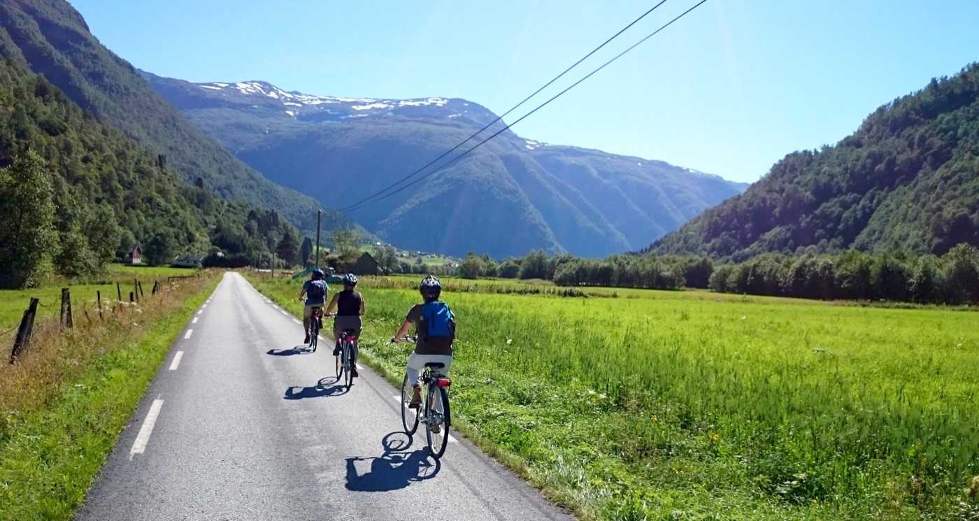 Three cyclists riding away up a narrow tarmac road between hay meadows on a green valley floor, a power line crossing overhead and snow still lying on the mountain shoulder at the head of the valley
