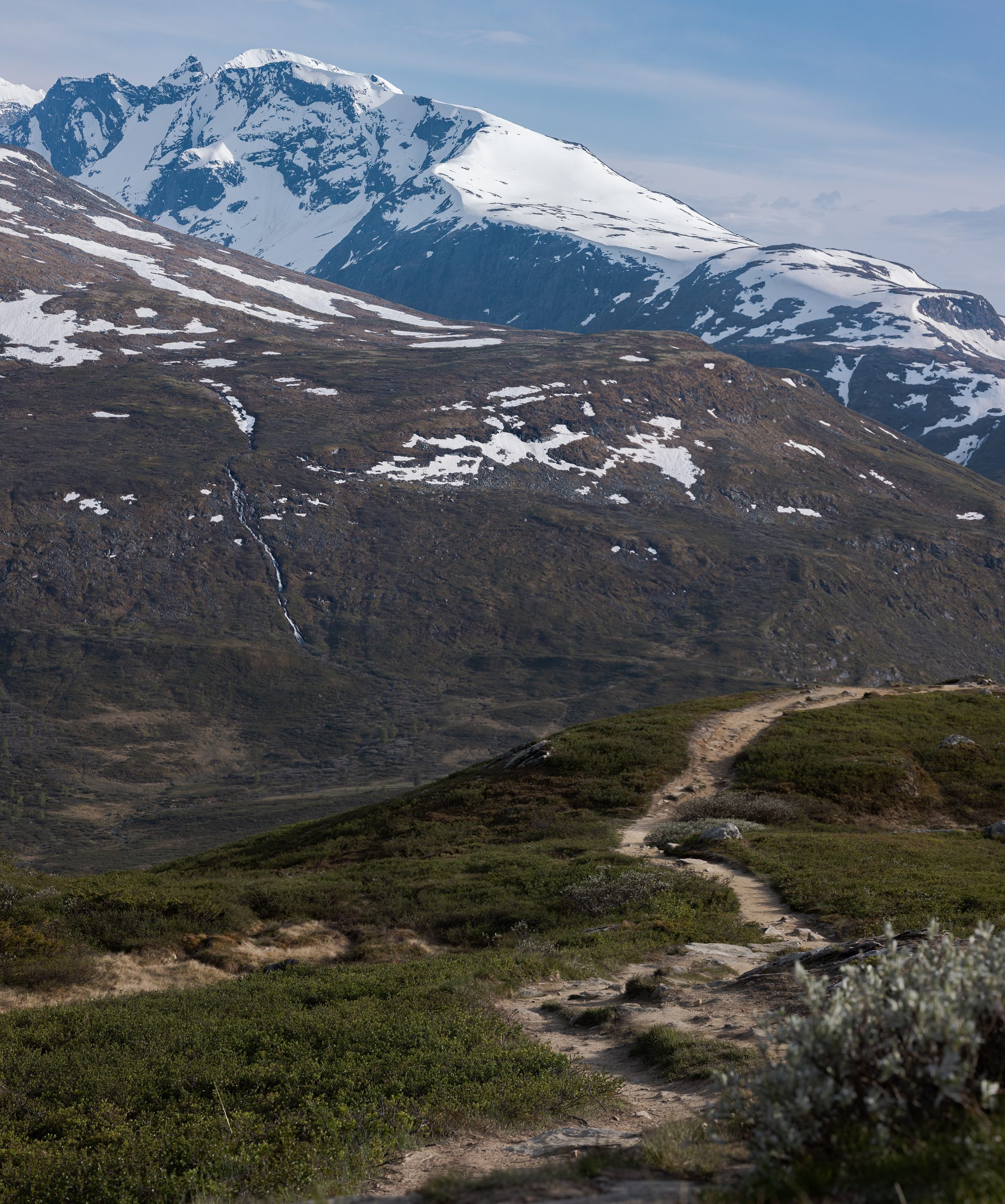 A worn path crossing a green mountainside towards a snow-covered peak, with meltwater streaks on the slope opposite