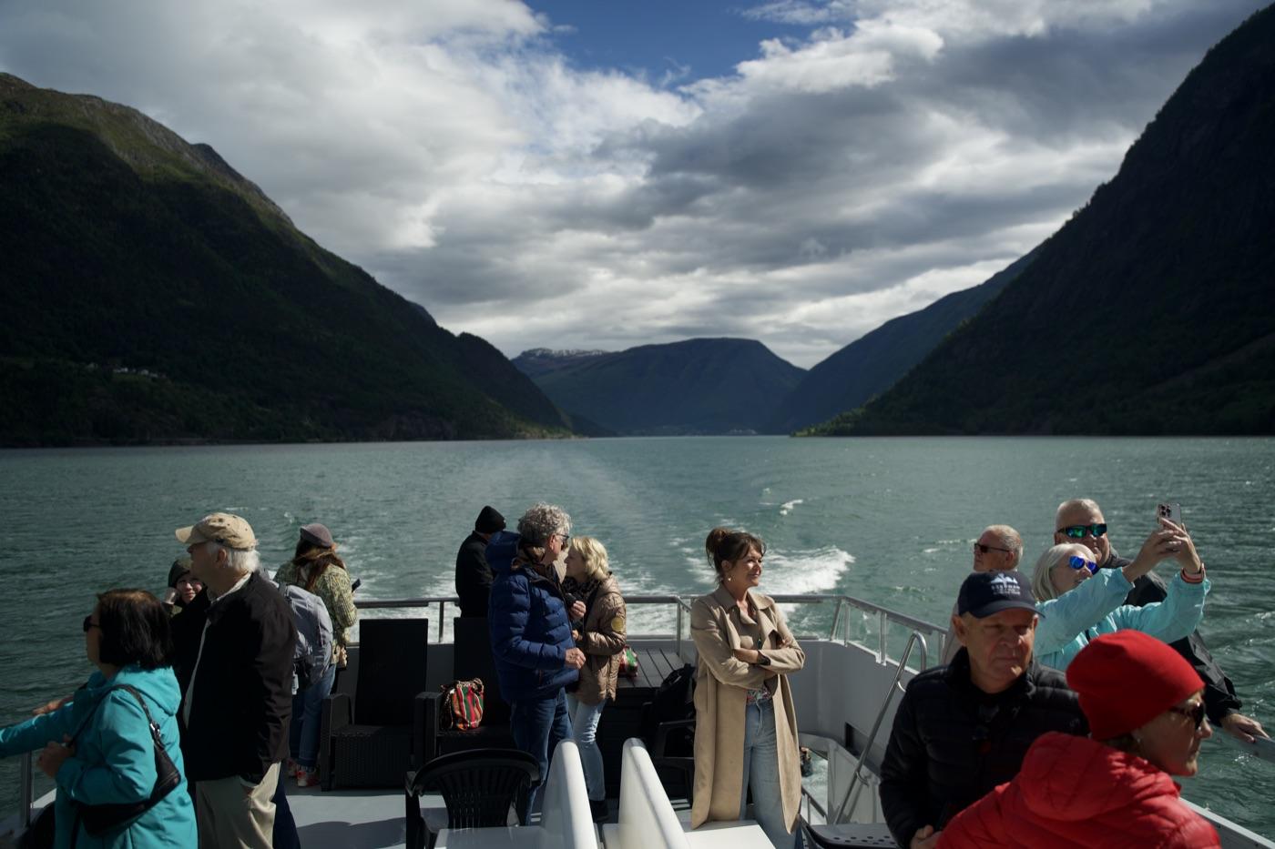 Guests standing and sitting on the open aft deck of the catamaran as she runs out the fjord, the wake spreading behind her and the water opening between two steep green mountainsides towards a distant snow-streaked ridge