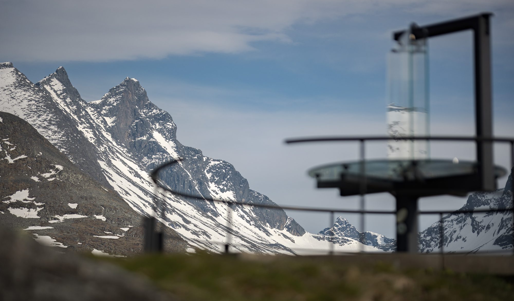 The glass panorama marker at Oscarshaug against broken cloud, with the sharp Hurrungane ridge rising to the left
